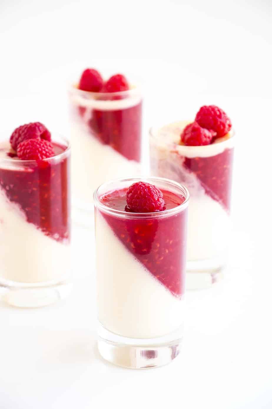 Four vanilla Raspberry Panna cottas in serving glasses topped with fresh raspberries.