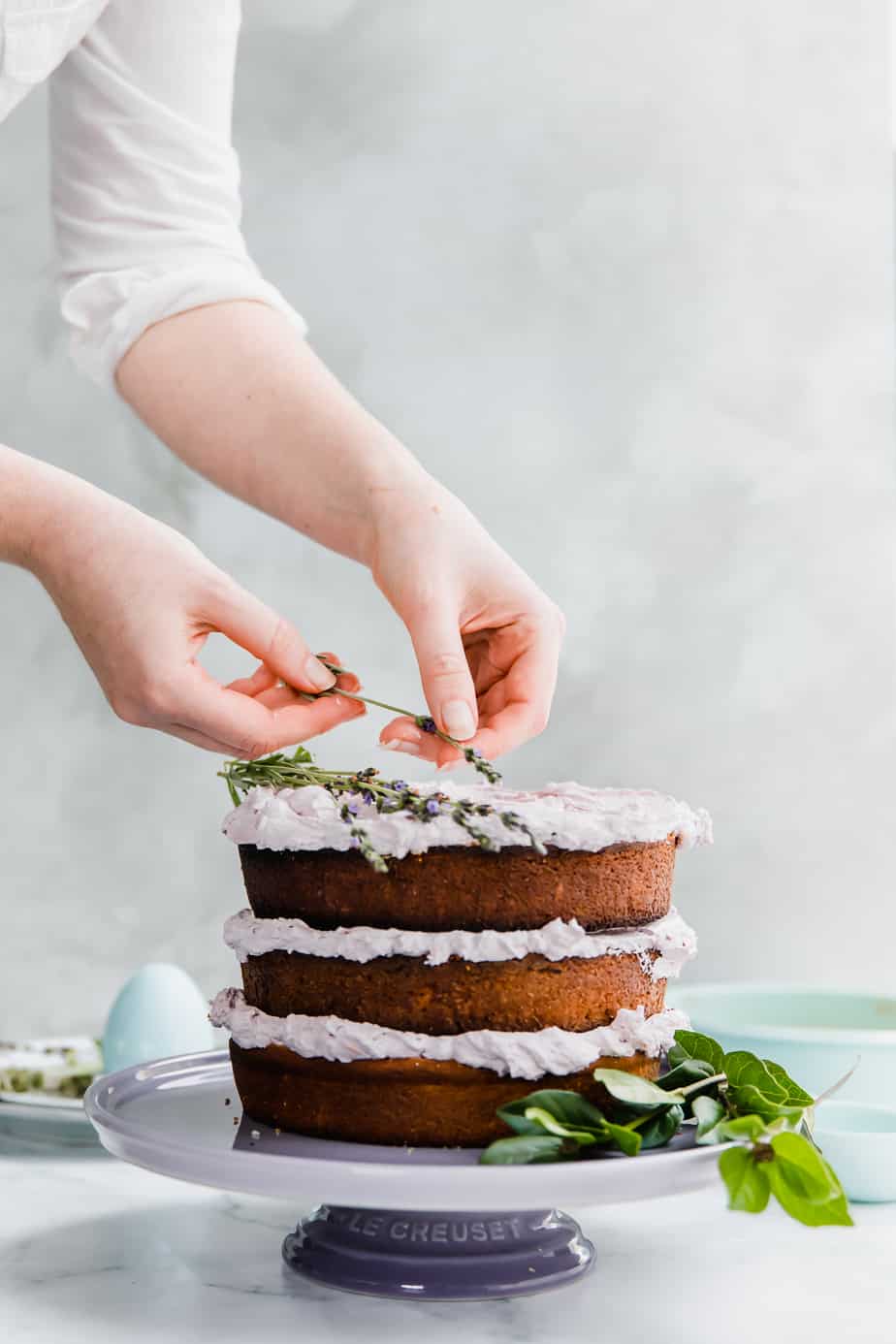 A hand placing fresh lavender on top of a layered cake on a cake stand.