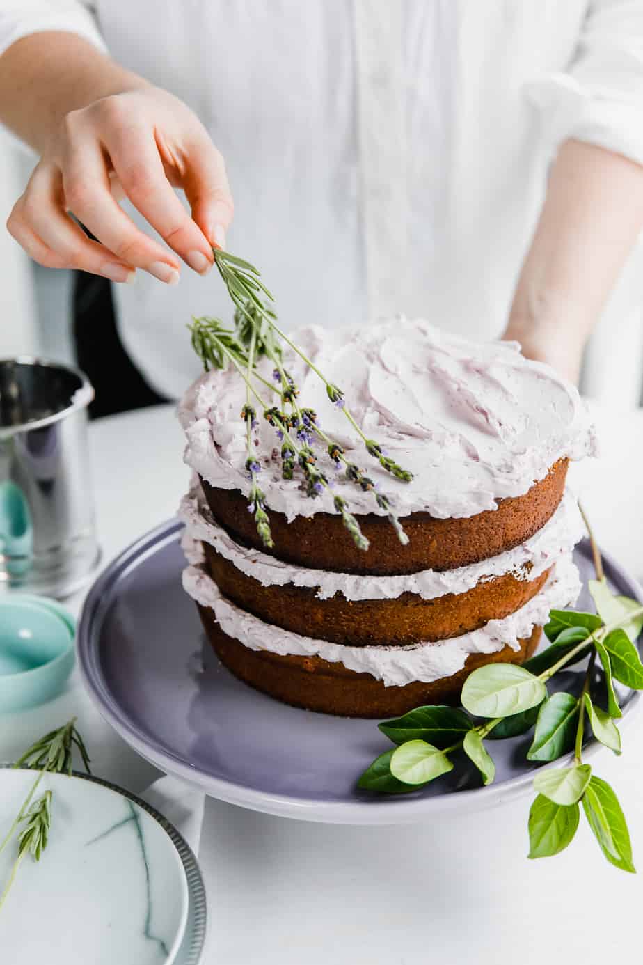 A person adding fresh lavender to an Earl Grey Lavender Cake on a purple cake stand.