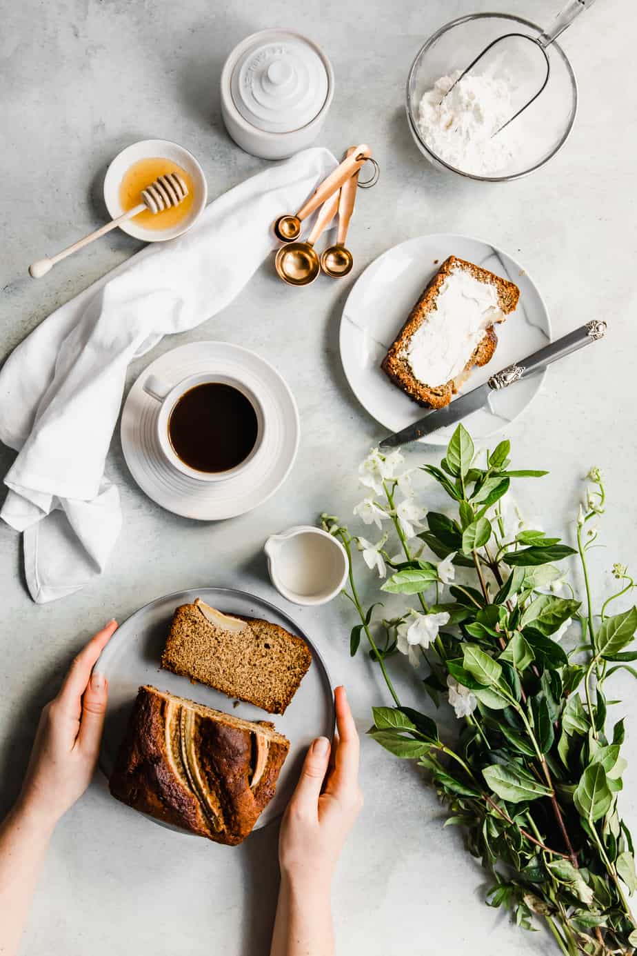 Banana bread with slices on a plate and cups of coffee and white flowers.