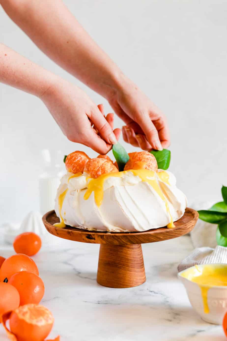 Hands topping a pavlova  on a wooden cake stand with clementine.