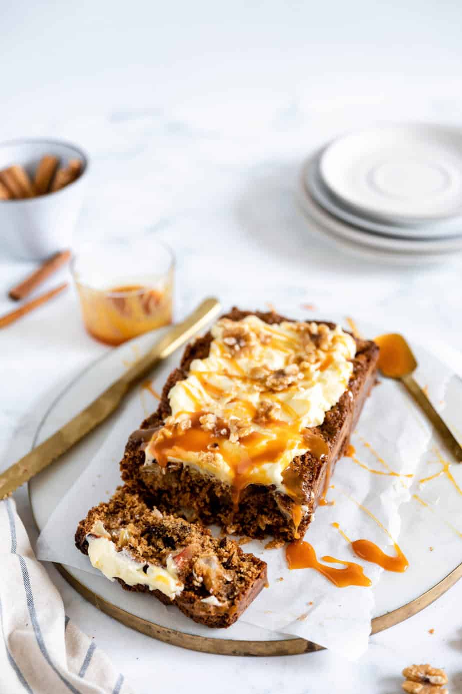 A apple and walnut loaf topped with creamy frosting and a drizzle of caramel sauce on a marble board.