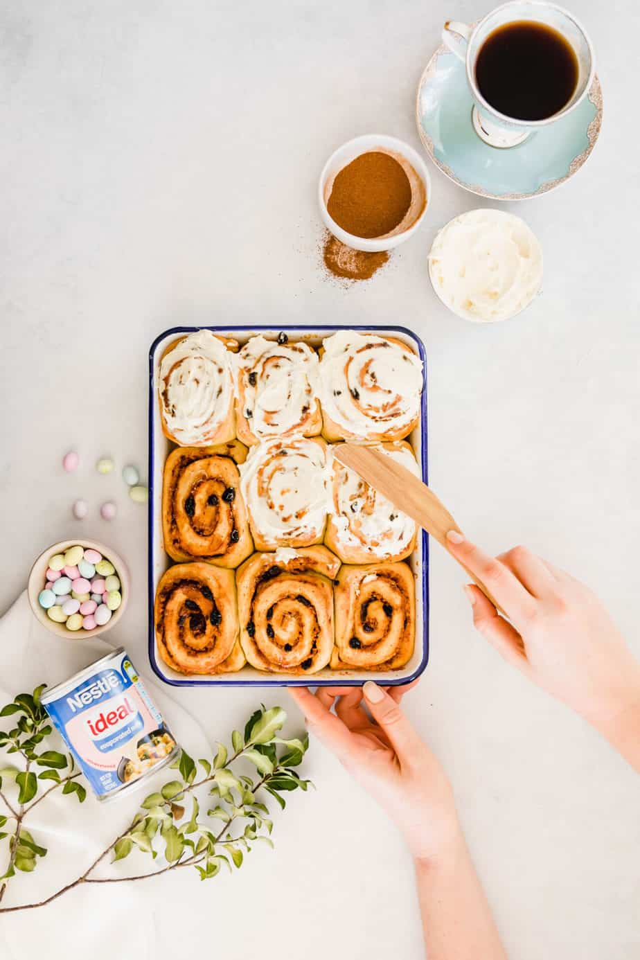A tray of cinnamon rolls being frosted with a wooden knife.