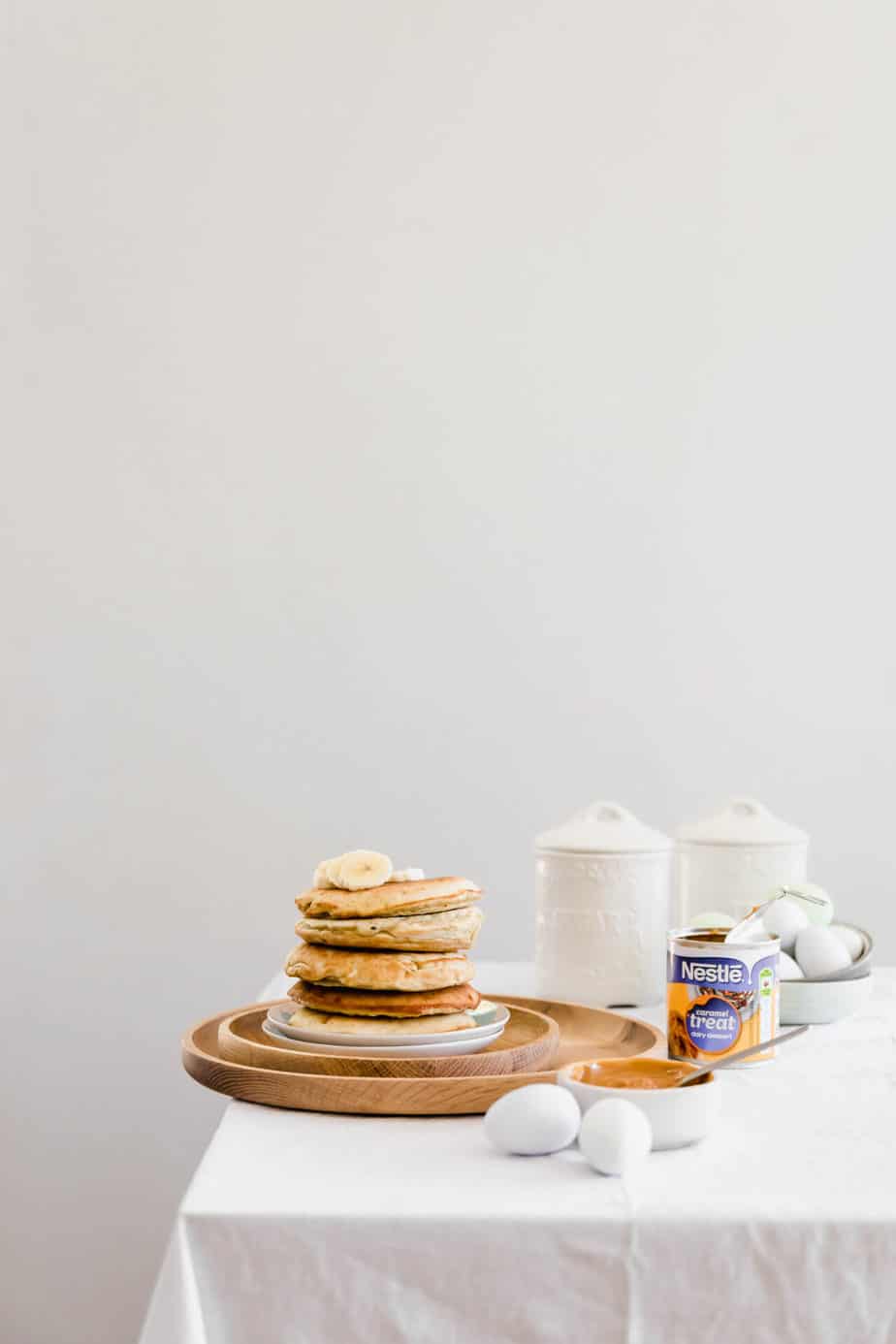A stack of pancakes on a wooden plate on the edge of a table.