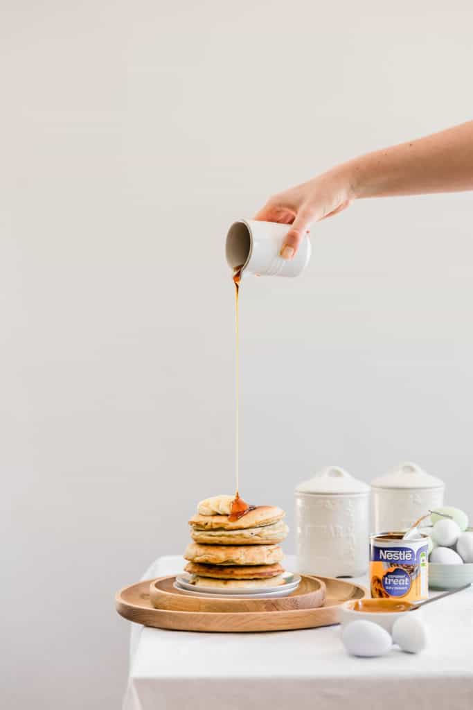 A jug pouring maple syrup over a stack of caramel pancakes with sliced bananas on top.