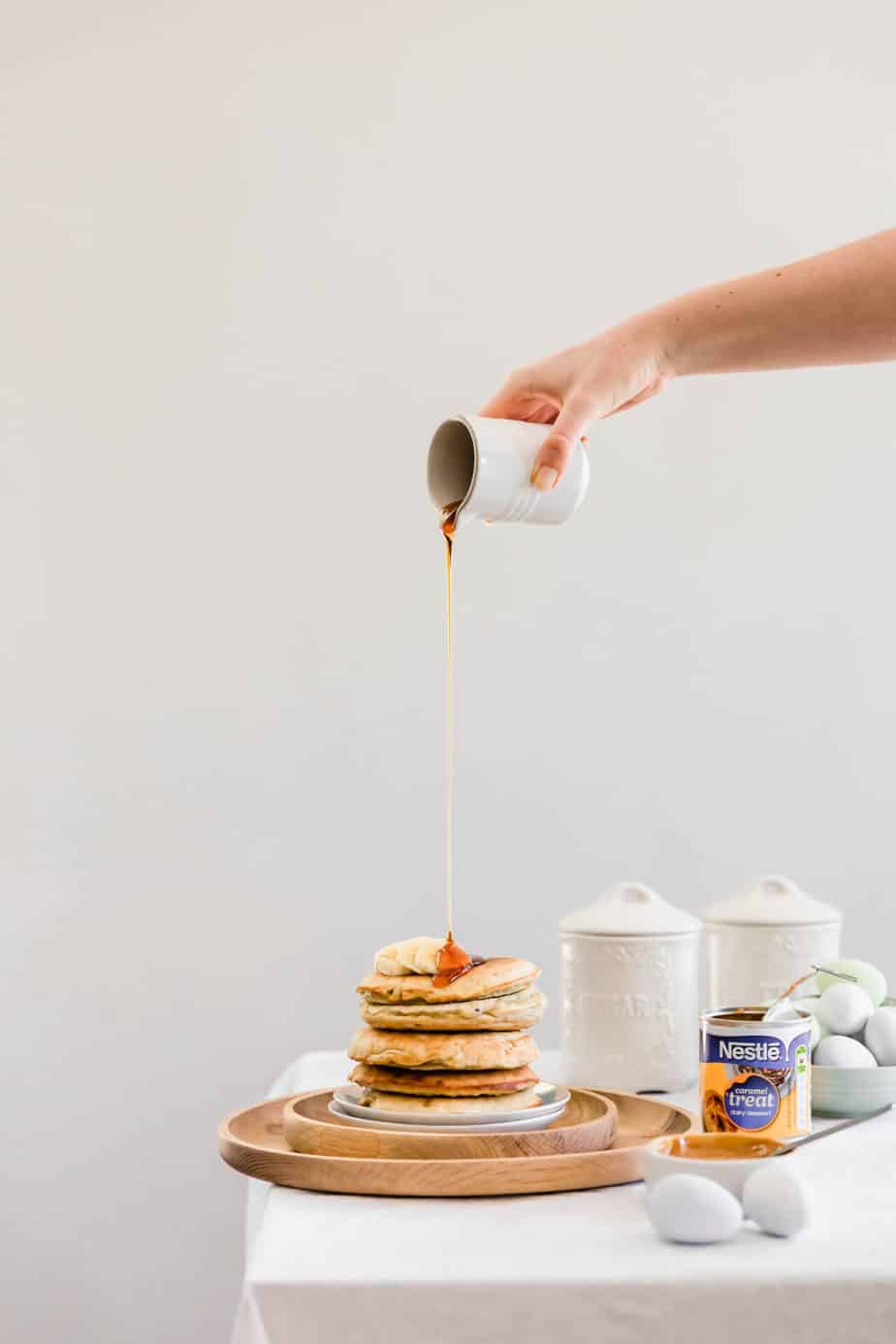 A jug pouring maple syrup over a stack of salted caramel pancakes with sliced bananas on top.