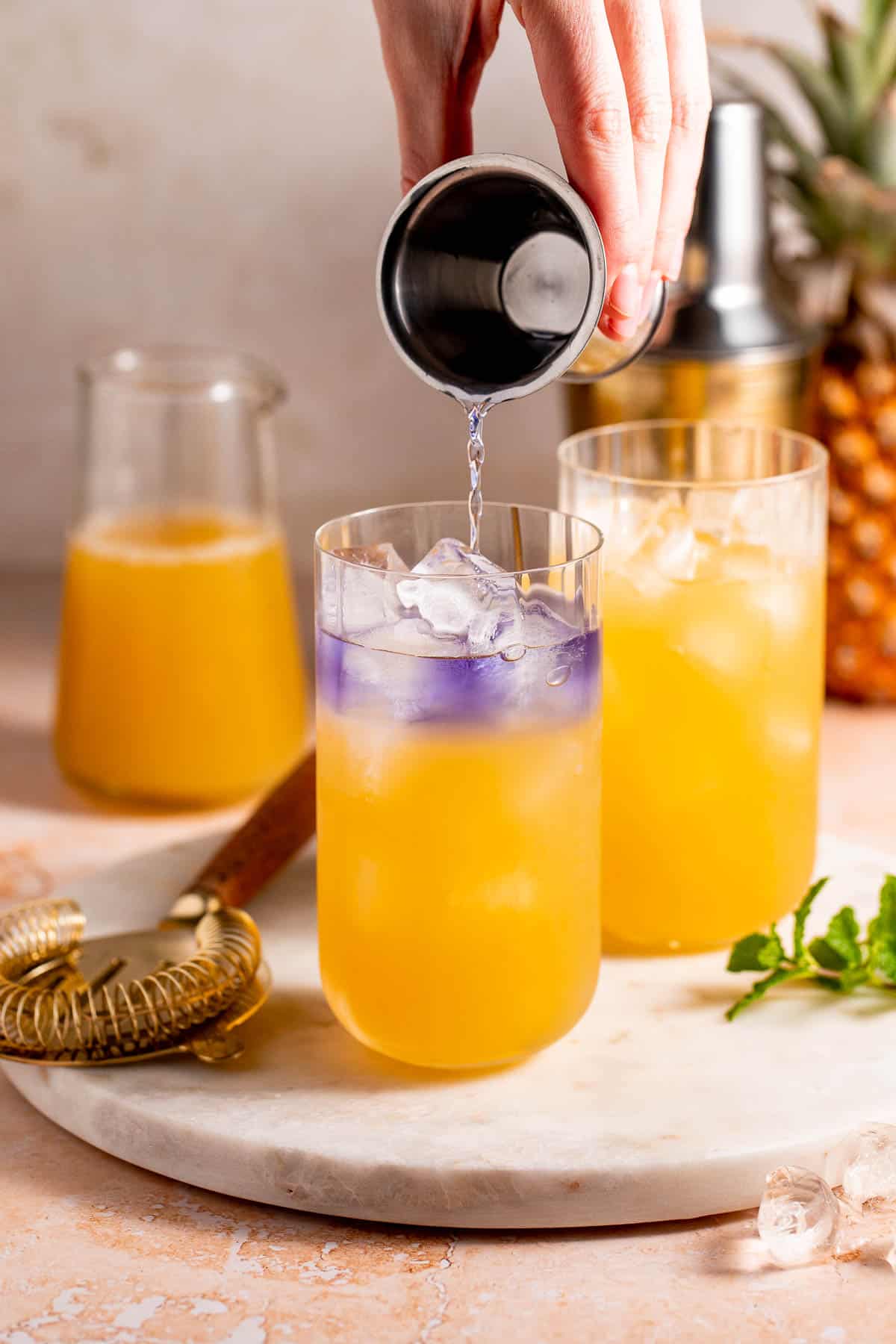 A purple liquid pouring slowly over an orange liquid in a serving glass.