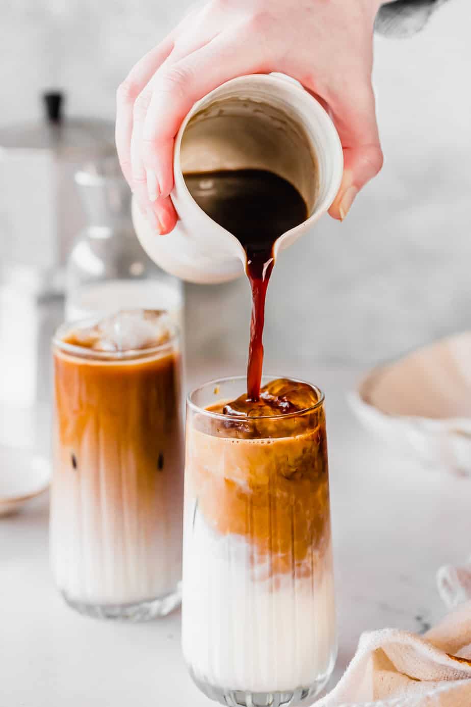 Coffee pouring from a jug into a glass with milk and ice with an iced coffee in the background.