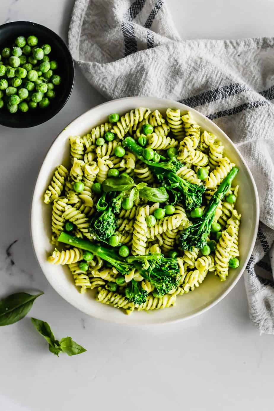 A bowl full of pesto pasta with broccoli and fresh peas and basil leaves.