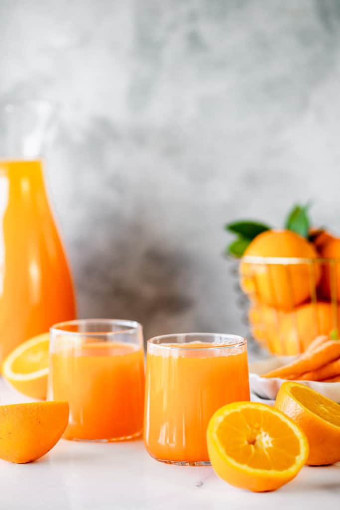 carrot and orange juice with basket of oranges and grey background