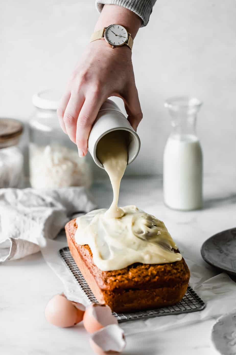 A jug pouring cream cheese frosting over a pumpkin bread loaf.