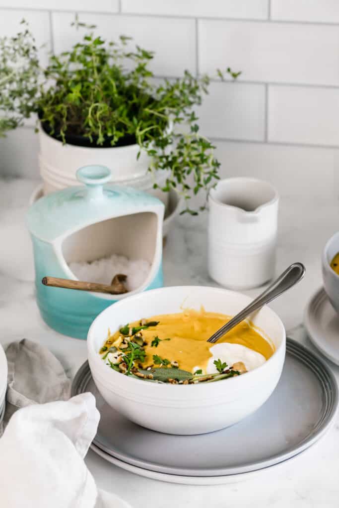 A bowl of creamy pumpkin lentil soup that is on a gray plate with a silver spoon.
