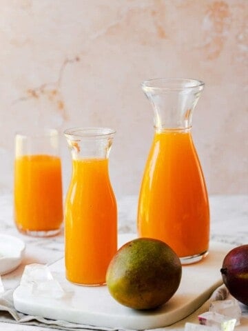 three jugs filled with carrot mango juice and a fresh whole mango on marble