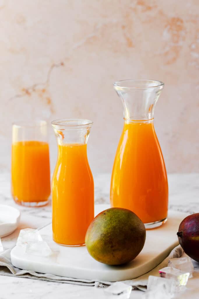 three jugs filled with carrot mango juice and a fresh whole mango on marble