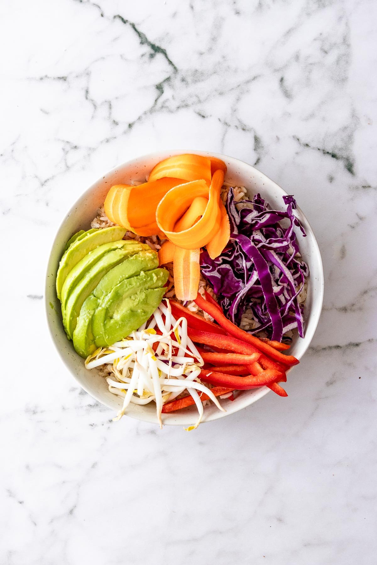 White bowl filled with rice, shredded cabbage, carrots, red pepper, bean sprouts, and avocado.