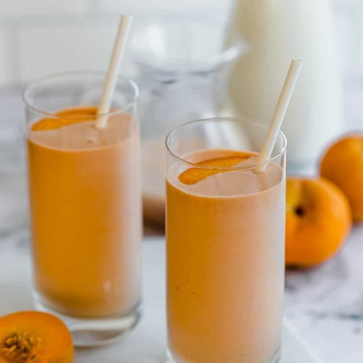 banana peach smoothie in tall glasses with straws and fresh whole peaches