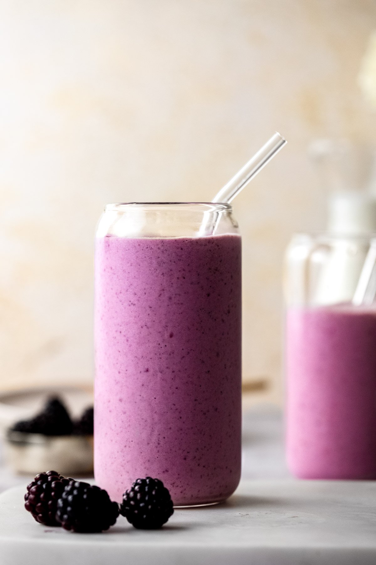 A glass filled with a purple smoothie and with a glass straw surrounded with fresh blackberries.