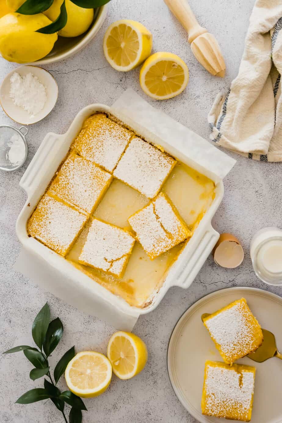 Lemon bars in a white dish with fresh lemons and powdered sugar.