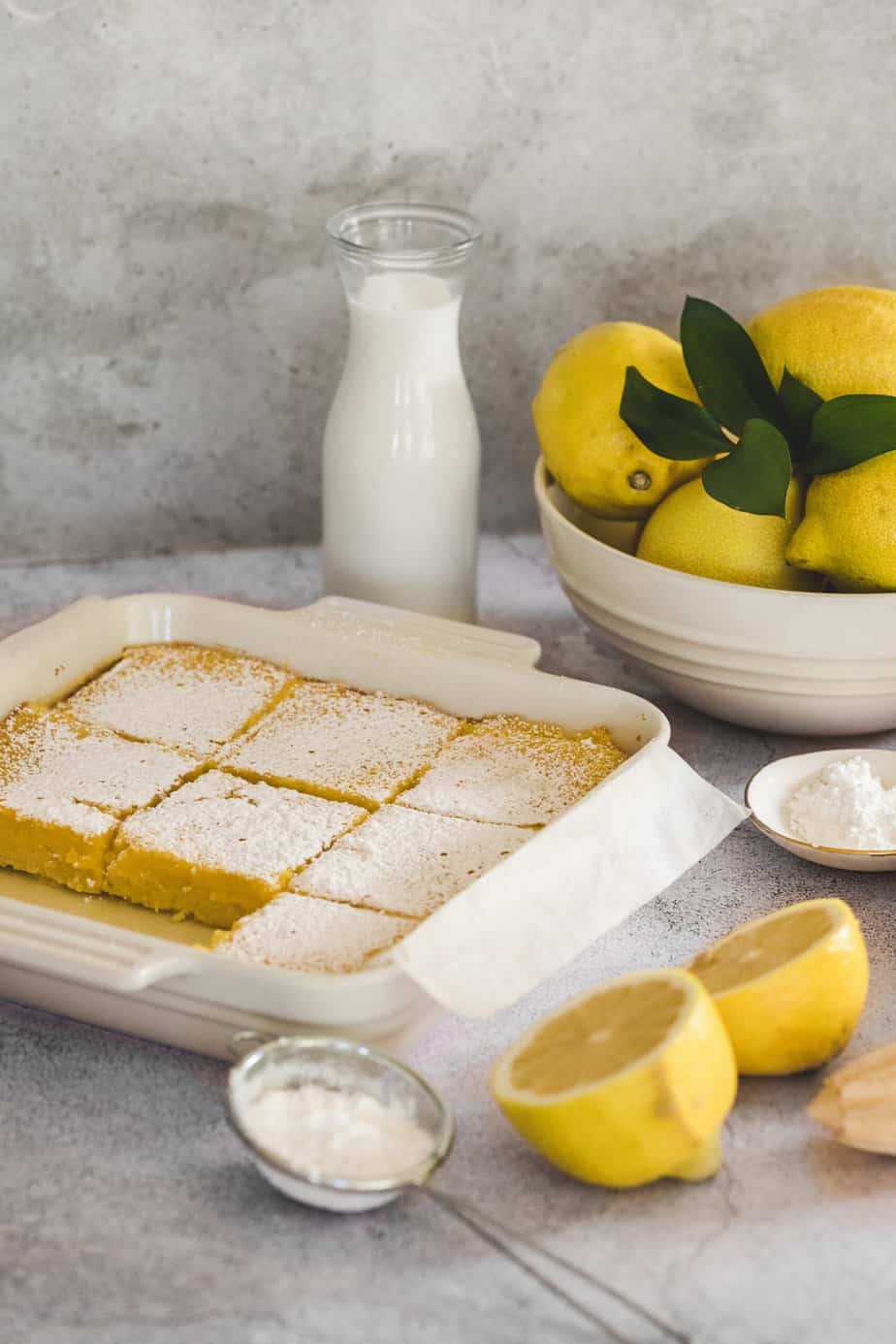 A dish of baked lemon bars with fresh lemons and powdered sugar.