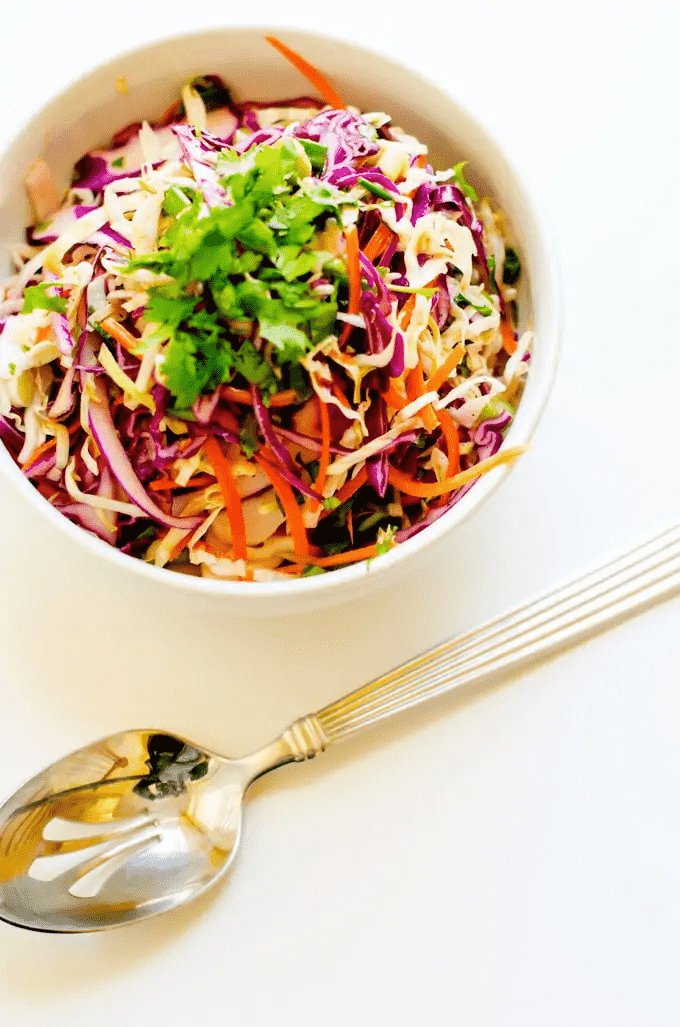 A rainbow slaw in a white bowl with a silver spoon is what to serve this summer.