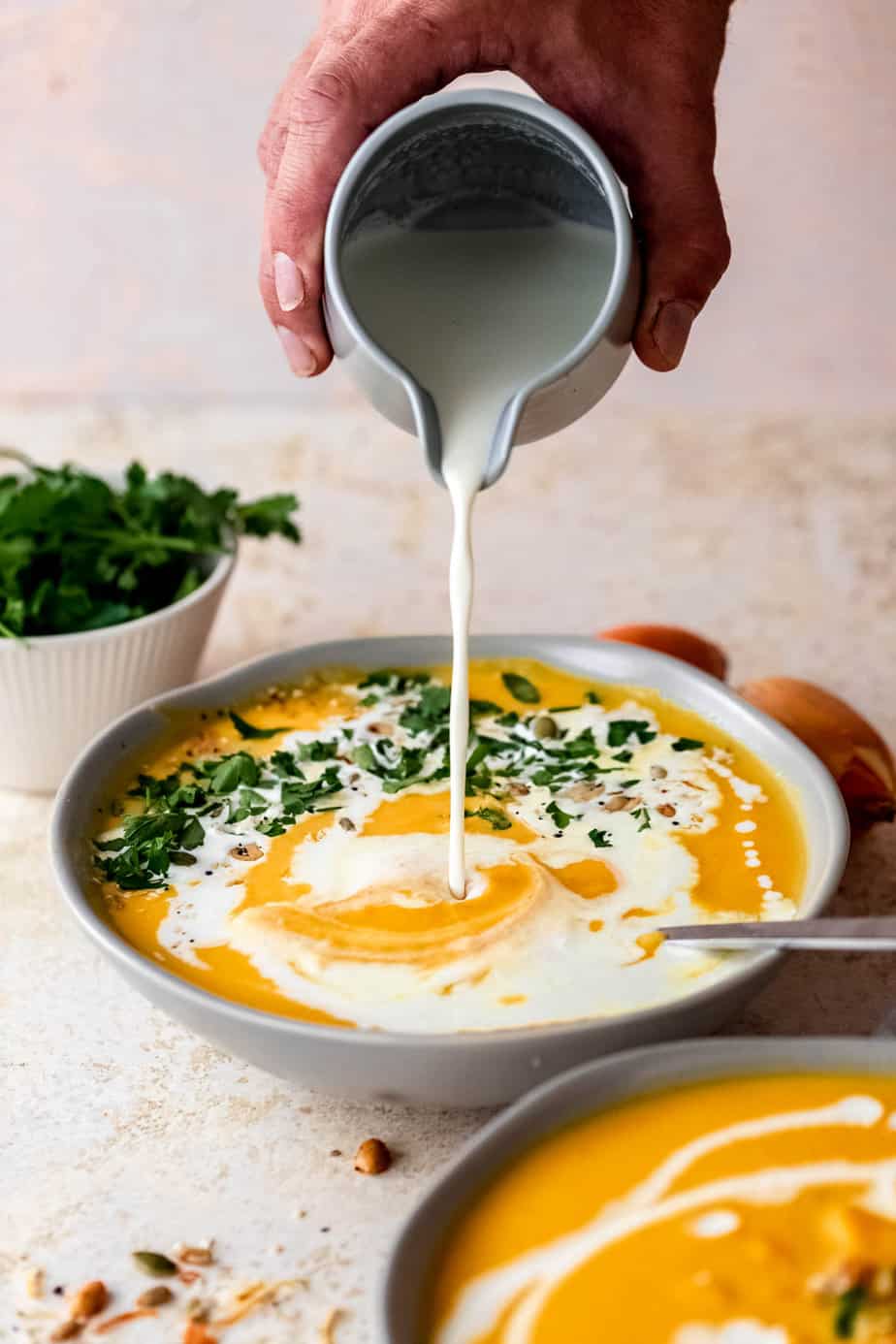 A jug pouring cream over a bowl of pumpkin soup and fresh herbs.