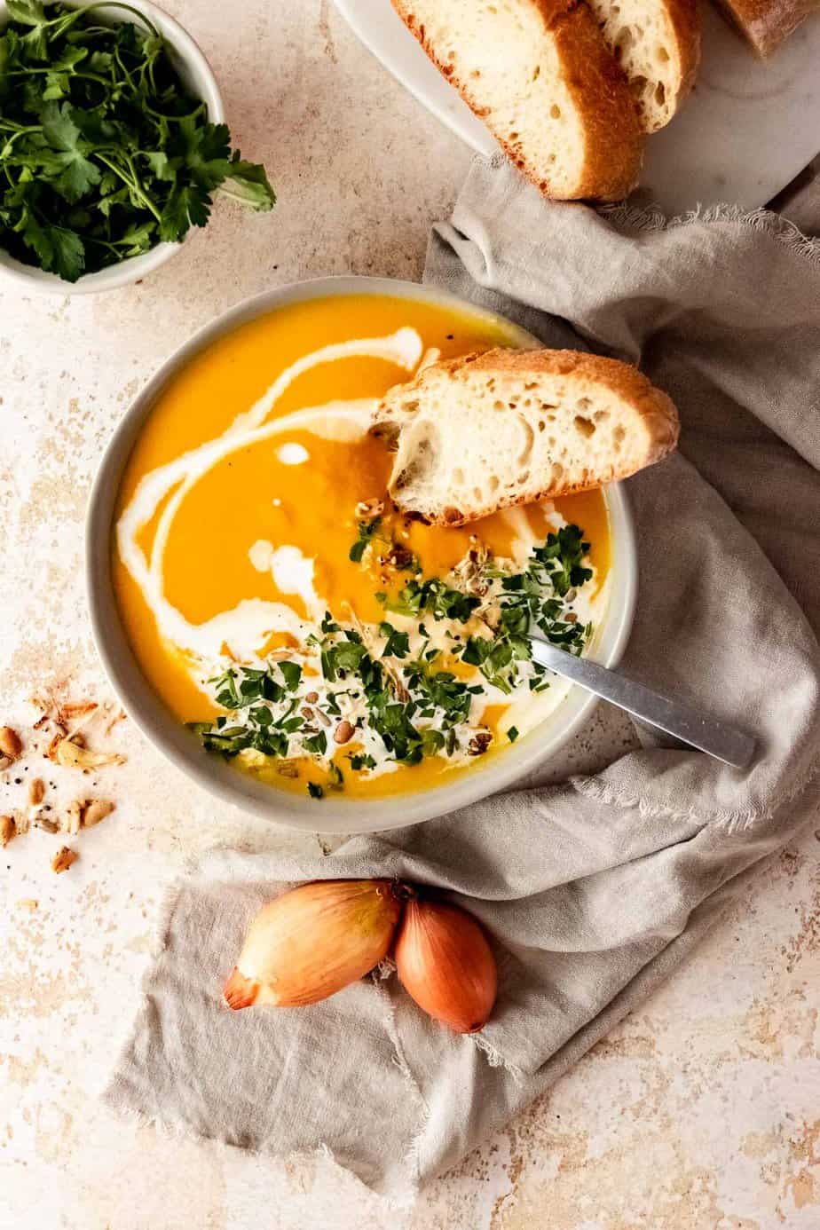 A bowl of pumpkin soup with a drizzle of cream and fresh herbs on top served with a slice of bread.