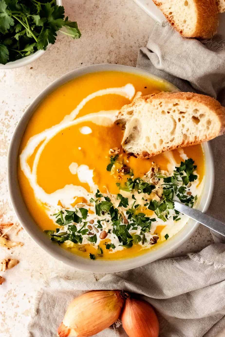 A large bowl of pumpkin soup with a spoon. The soup is garnished with fresh herbs, bread, cream, and seeds.