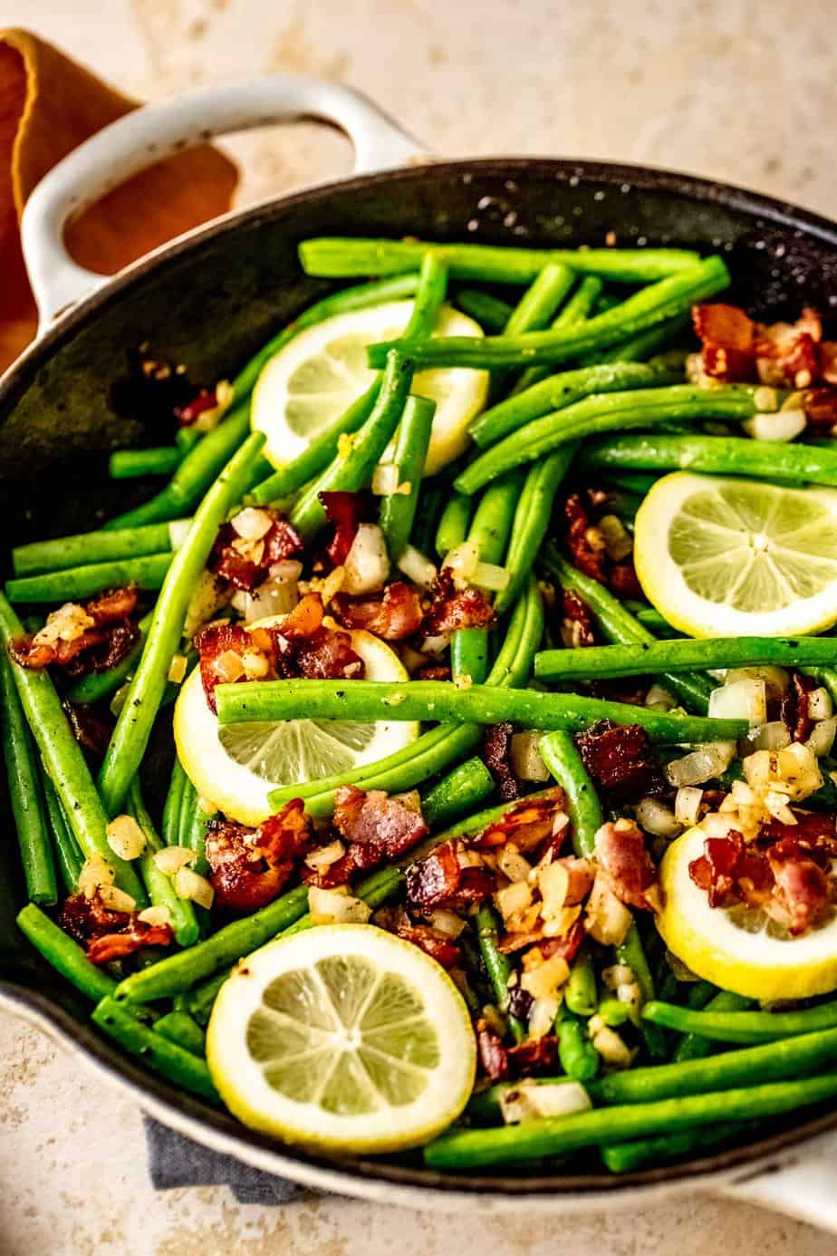A skillet filled with green beans and bacon topped with slices of fresh lemon