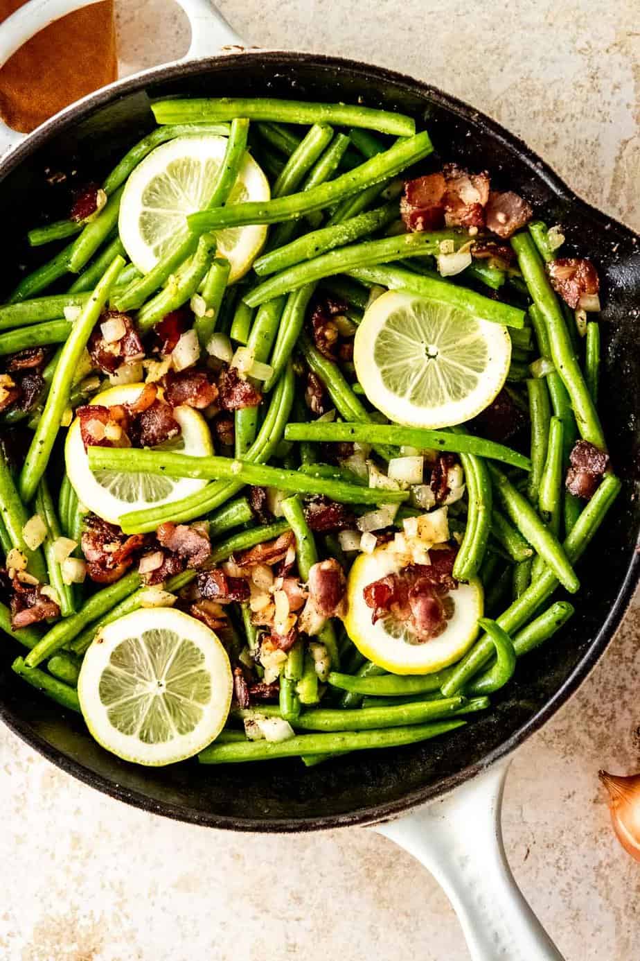 A skillet with green beans, onion, and bacon garnished with lemon slices.