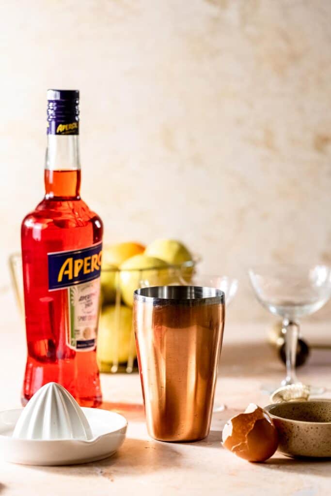 aperol sour bottle with rose gold cocktail shaker and white lemon squeezer