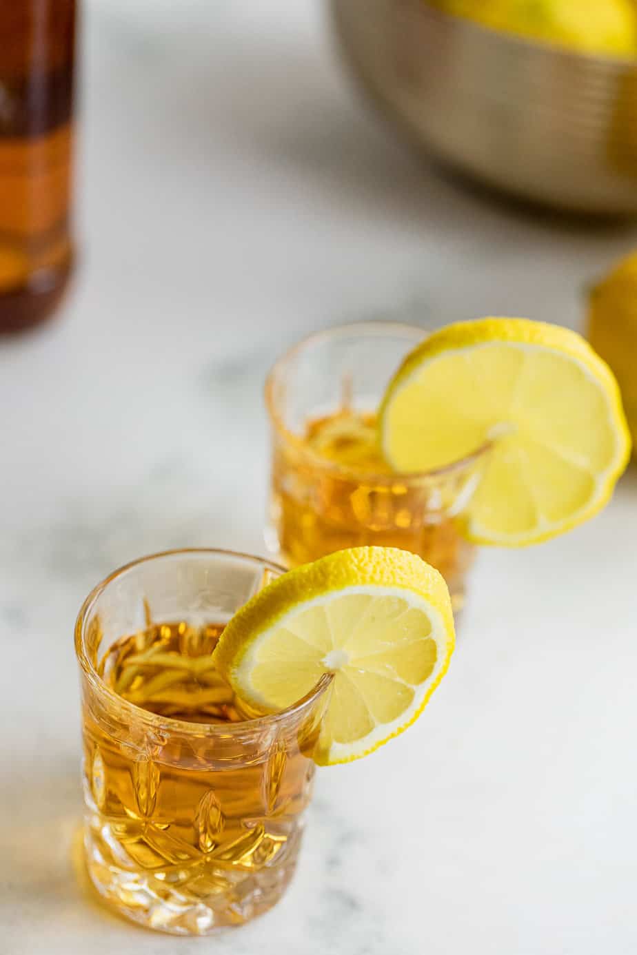 two whiskey shots with slices of lemon and bottle of whiskey in the background