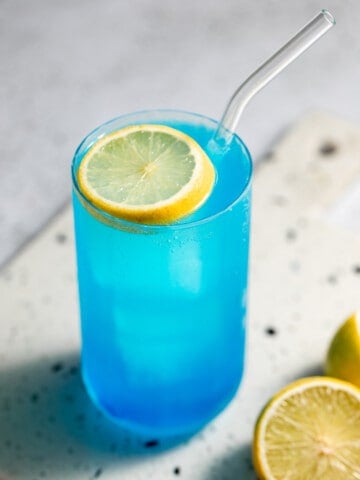 blue drink in tall glass with lemon slice as garnish and glass straw