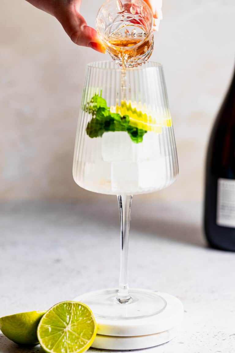 Elderflower liqueur being poured into a glass filled with ice, lime slices, and mint.