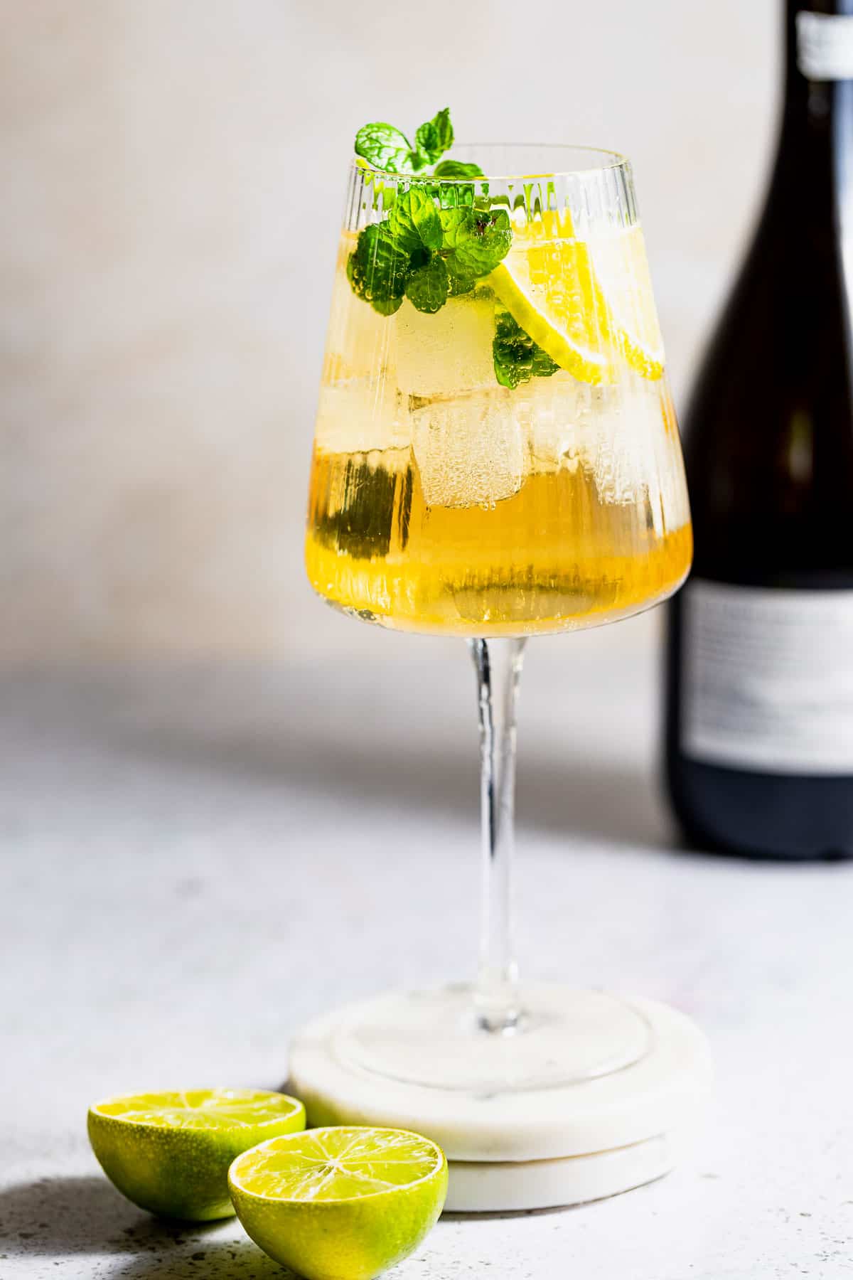A Hugo Spritz cocktail with ice, mint, and lemon slices and a bottle of prosecco in the background.