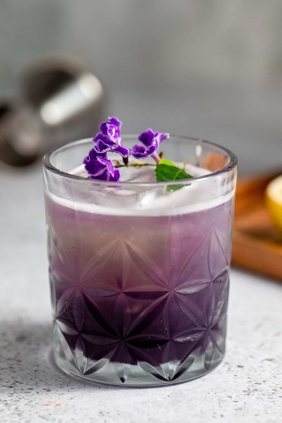 one glass of purple rain cocktail with ice and purple flowers